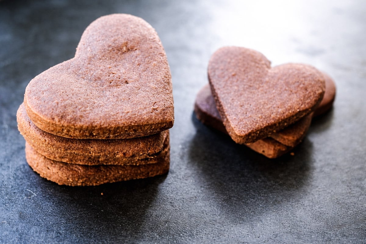 A stack of heart-shaped chocolate cookies and espresso shortbreads sits on a dark surface, with three more heart-shaped cookies lying nearby.