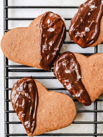 Four heart-shaped Espresso Shortbreads on a cooling rack-three are partially dipped in chocolate and sprinkled with coarse sea salt, while one cookie is plain without chocolate or salt.