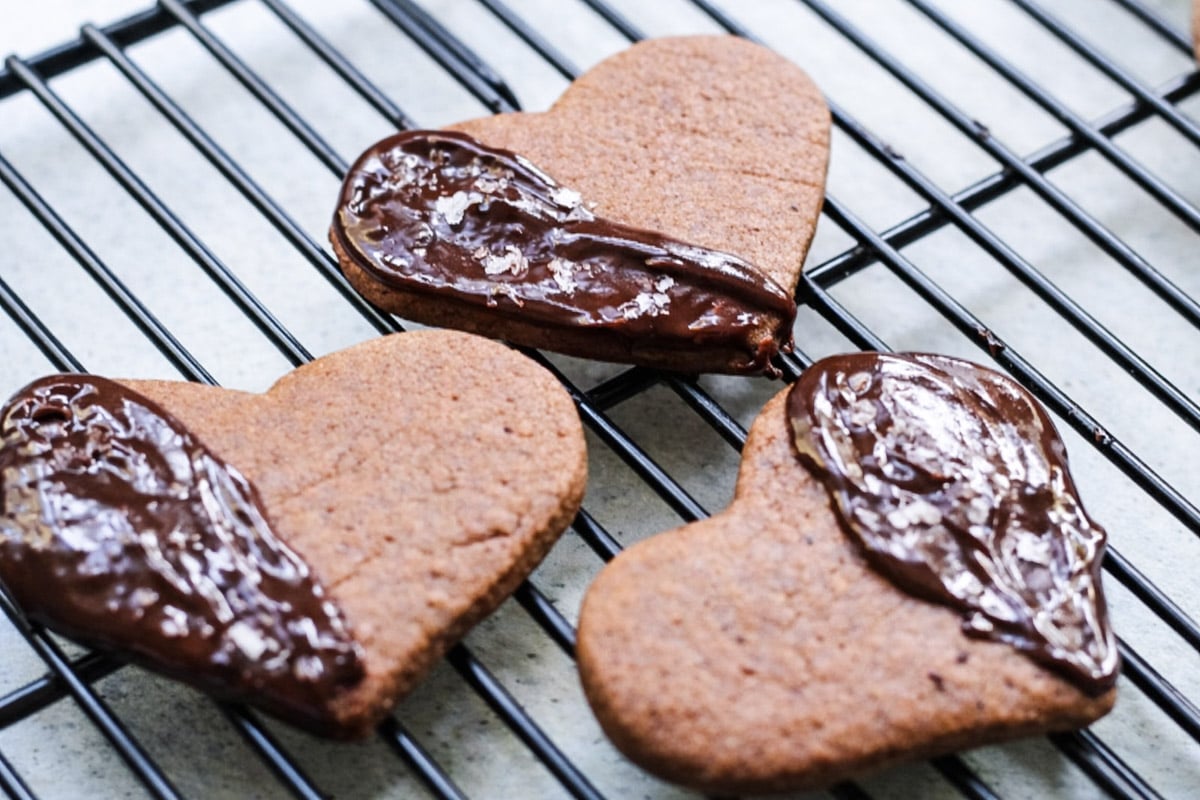 Three heart-shaped Espresso Shortbreads, each with one half dipped in chocolate, rest elegantly on a black wire cooling rack.