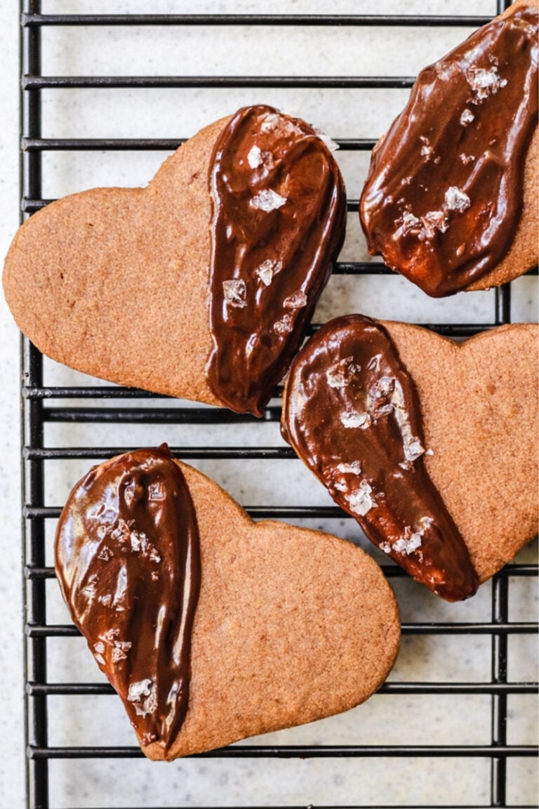 Four heart-shaped Espresso Shortbreads on a cooling rack—three are partially dipped in chocolate and sprinkled with coarse sea salt, while one cookie is plain without chocolate or salt.