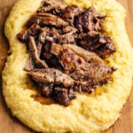 Shredded braised beef served on a bed of creamy yellow polenta, reminiscent of classic Slow Roasted Italian Pork with Polenta, arranged beautifully on a wooden cutting board.