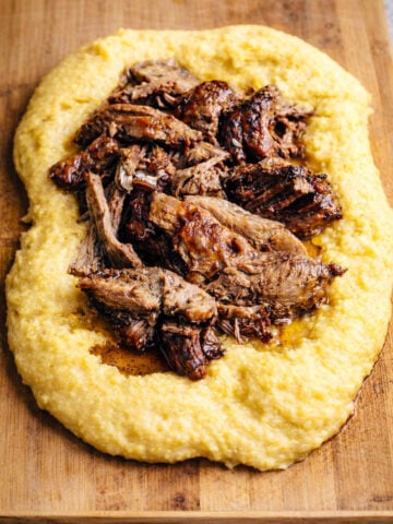 Shredded braised beef served on a bed of creamy yellow polenta, reminiscent of classic Slow Roasted Italian Pork with Polenta, arranged beautifully on a wooden cutting board.