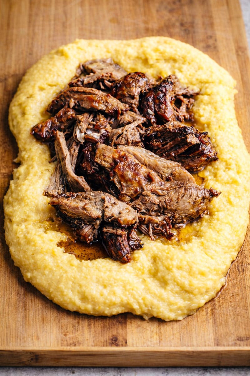 Shredded braised beef served on a bed of creamy yellow polenta, reminiscent of classic Slow Roasted Italian Pork with Polenta, arranged beautifully on a wooden cutting board.