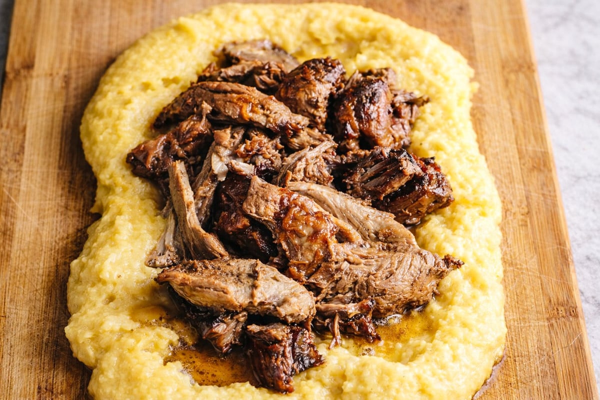 Shredded, juicy braised beef piled on top of a creamy bed of yellow polenta, reminiscent of Slow Roasted Italian Pork with Polenta, served on a wooden cutting board.