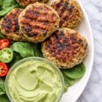 Four grilled Spinach Chicken Burgers rest on a bed of spinach, served with sliced red and green peppers, a pickle spear, and a small bowl of creamy avocado dip on a white plate over marble.