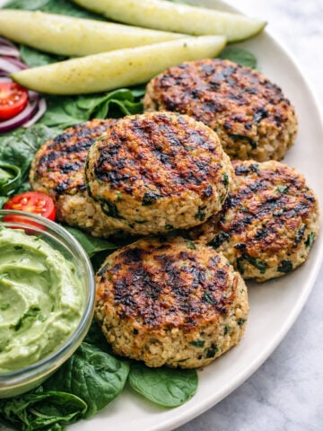 Four Spinach Chicken Burgers are served on a plate with fresh spinach, sliced cherry tomatoes, cucumber spears, red onion rings, and a small bowl of creamy green avocado dip.