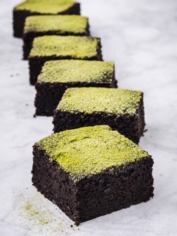 Six Vegan Matcha Brownies are arranged in a diagonal line on a marble surface, each topped with a vibrant green powder, likely matcha.
