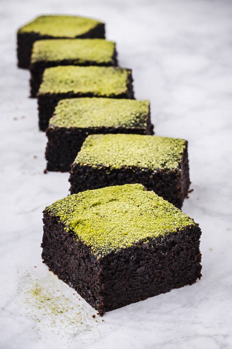 Six Vegan Matcha Brownies are arranged in a diagonal line on a marble surface, each topped with a vibrant green powder, likely matcha.