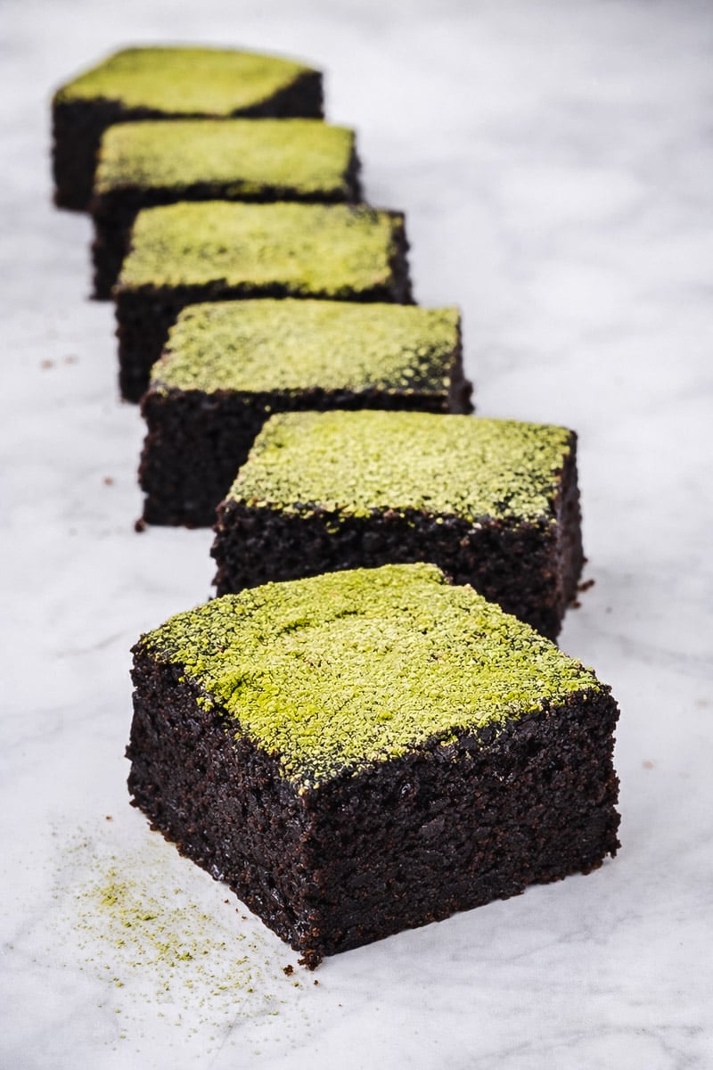 Six Vegan Matcha Brownies are arranged in a diagonal line on a marble surface, each topped with a vibrant green powder, likely matcha.