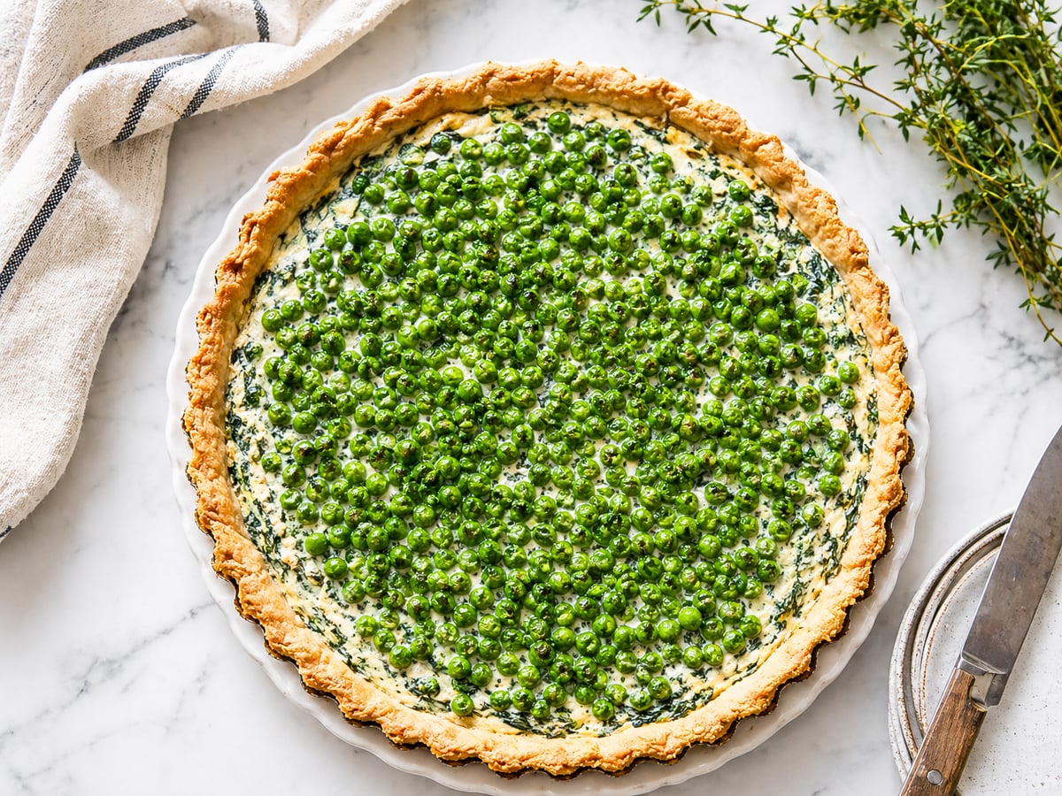 A freshly baked Spring Pea and Ricotta Tart with a golden crust, filled with green peas and creamy filling, sits on a marble surface next to a kitchen towel, knife, and sprigs of fresh herbs.