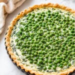 A freshly baked Spring Pea and Ricotta Tart with a flaky crust, filled with creamy spinach and cheese, and topped with vibrant green peas, sits on a marble surface next to a beige cloth and a green leafy sprig.