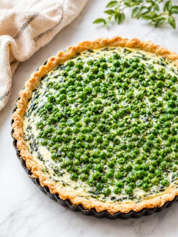 A freshly baked Spring Pea and Ricotta Tart with a flaky crust, filled with creamy spinach and cheese, and topped with vibrant green peas, sits on a marble surface next to a beige cloth and a green leafy sprig.