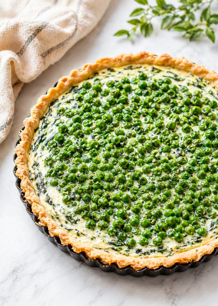 A freshly baked Spring Pea and Ricotta Tart with a flaky crust, filled with creamy spinach and cheese, and topped with vibrant green peas, sits on a marble surface next to a beige cloth and a green leafy sprig.
