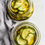 Two glass jars filled with sliced pickles in brine sit on a light surface next to a gray cloth. The pickles, inspired by Easy Marinated Zucchini, are seasoned with visible herbs and spices for extra flavor.