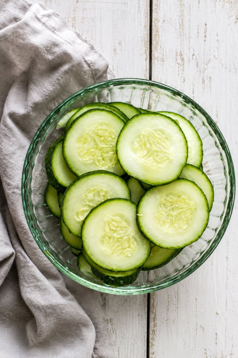 A glass bowl filled with fresh cucumber slices and Easy Marinated Zucchini sits on a white wooden surface next to a light gray cloth napkin.
