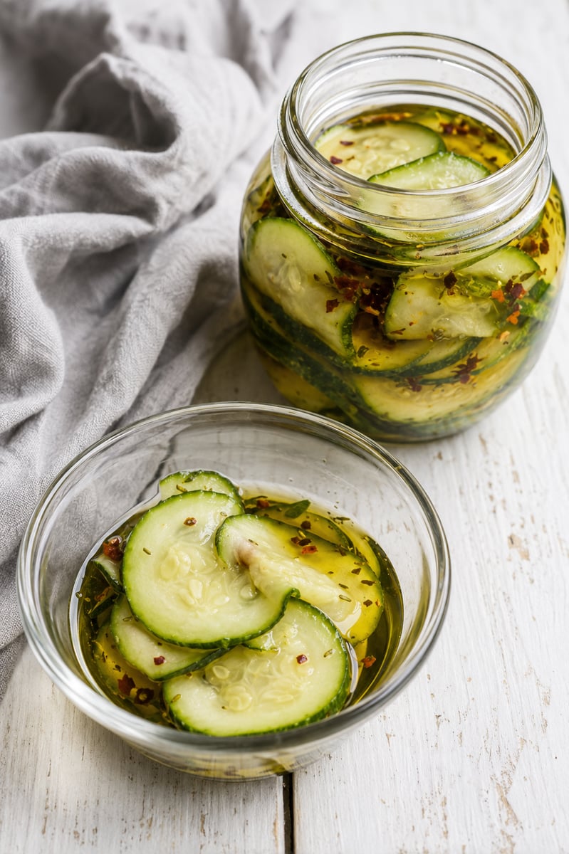 Glass jar and bowl filled with cucumber slices and Easy Marinated Zucchini in oil and spices, sitting on a white wooden surface with a light gray cloth nearby.