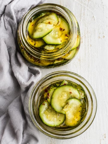 Two glass jars filled with sliced pickles in brine sit on a light surface next to a gray cloth. The pickles, inspired by Easy Marinated Zucchini, are seasoned with visible herbs and spices for extra flavor.