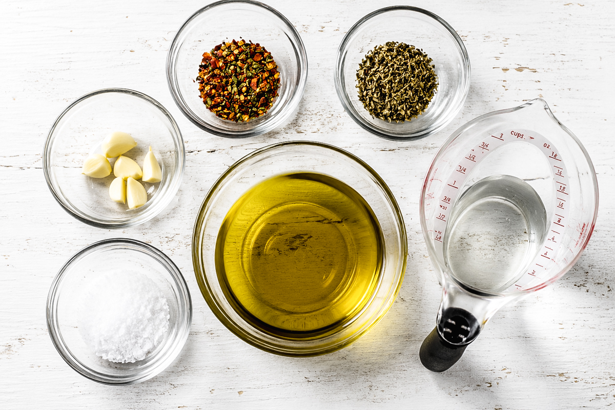Six glass containers hold olive oil, water, salt, garlic cloves, dried chili flakes, and dried herbs-perfect ingredients for Easy Marinated Zucchini-arranged neatly on a white wooden surface.