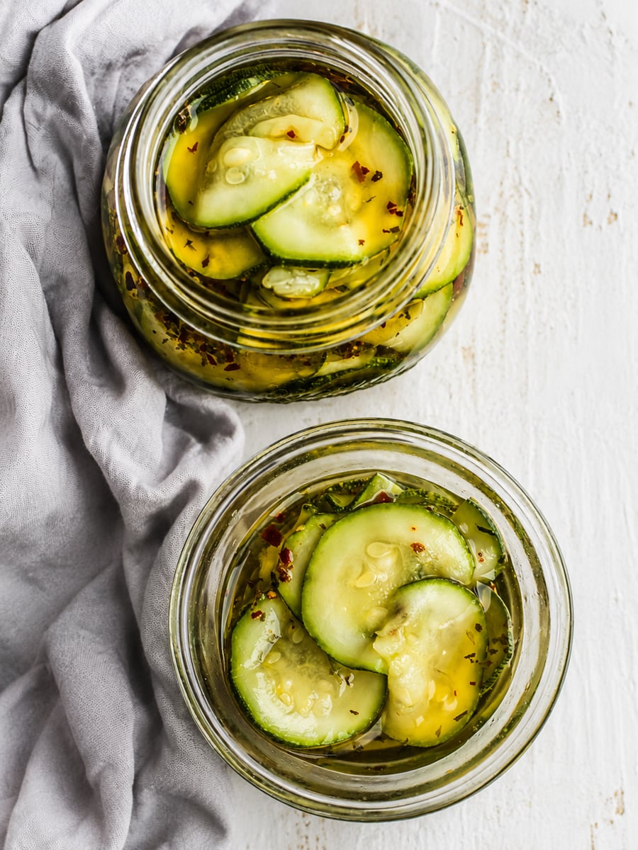 Two glass jars filled with sliced pickles in brine sit on a light surface next to a gray cloth. The pickles, inspired by Easy Marinated Zucchini, are seasoned with visible herbs and spices for extra flavor.