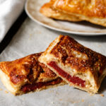 Two flaky pastries with golden crusts are filled with a bright red, jelly-like filling, capturing the essence of Pastelitos de Guayaba-classic Cuban Guava Pastries. One pastry is cut in half, revealing the luscious filling inside. More pastries sit on a plate in the background.