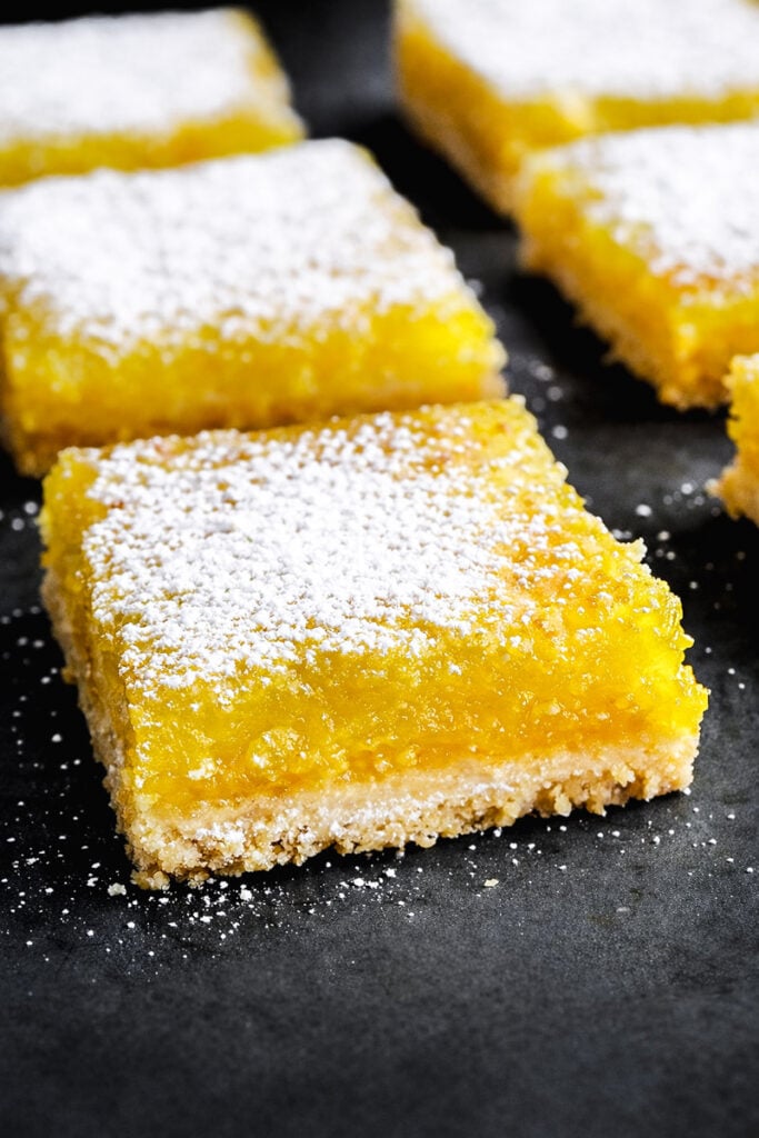 Close-up of Easy Lemon Bars with a golden, crumbly crust topped with bright yellow lemon filling and a dusting of powdered sugar, arranged on a dark surface.