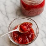 A glass jar and a small glass bowl filled with red strawberry jam sit on a white marble surface. A spoon rests in the bowl, just as it would for serving an easy plum jam.
