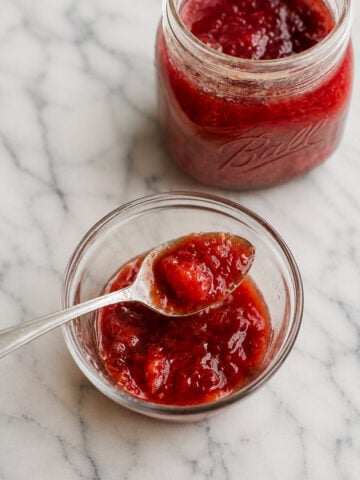 A glass jar and a small glass bowl filled with red strawberry jam sit on a white marble surface. A spoon rests in the bowl, just as it would for serving an easy plum jam.