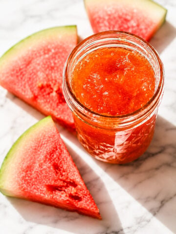 A glass jar filled with bright red Watermelon Rind Jam sits on a marble surface, surrounded by three fresh watermelon slices with green rinds. Sunlight highlights the vibrant colors and textures.