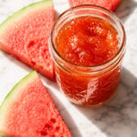 A glass jar filled with watermelon rind jam sits on a marble surface, surrounded by three triangular slices of fresh watermelon.