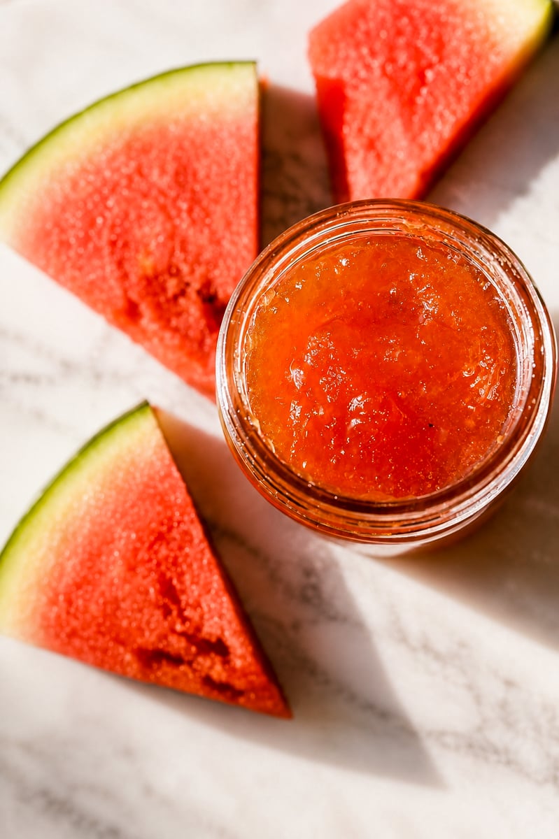 A jar of vibrant orange-red Watermelon Rind Jam sits on a marble surface, surrounded by three triangular slices of fresh watermelon with green rinds.
