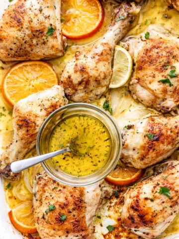 A sheet pan mojo chicken dish filled with roasted chicken pieces, orange and lemon slices, and fresh herbs, with a small bowl of herb-infused oil and a spoon in the center.