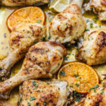 Sheet Pan Mojo Chicken drumsticks and thighs are seasoned with herbs, arranged on a tray with slices of orange and lemon, and garnished with chopped parsley.