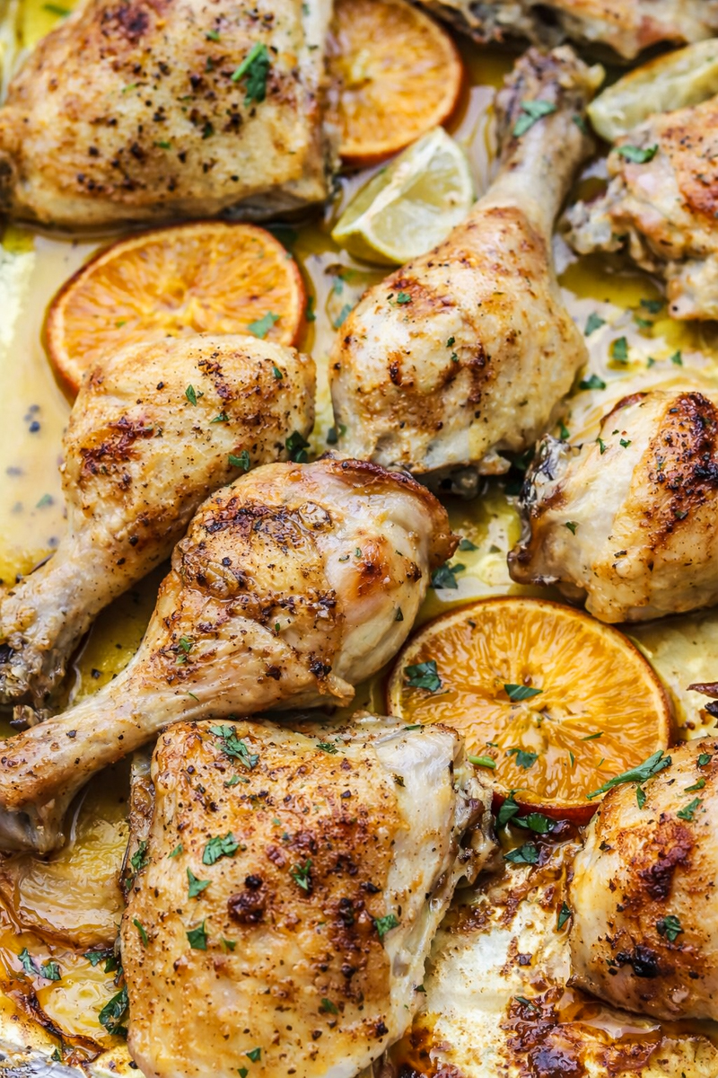 Sheet Pan Mojo Chicken drumsticks and thighs are seasoned with herbs, arranged on a tray with slices of orange and lemon, and garnished with chopped parsley.