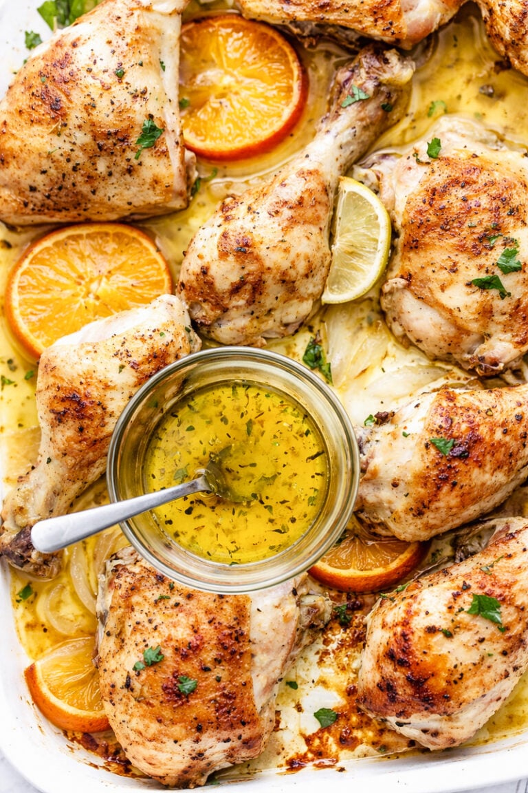 A sheet pan mojo chicken dish filled with roasted chicken pieces, orange and lemon slices, and fresh herbs, with a small bowl of herb-infused oil and a spoon in the center.