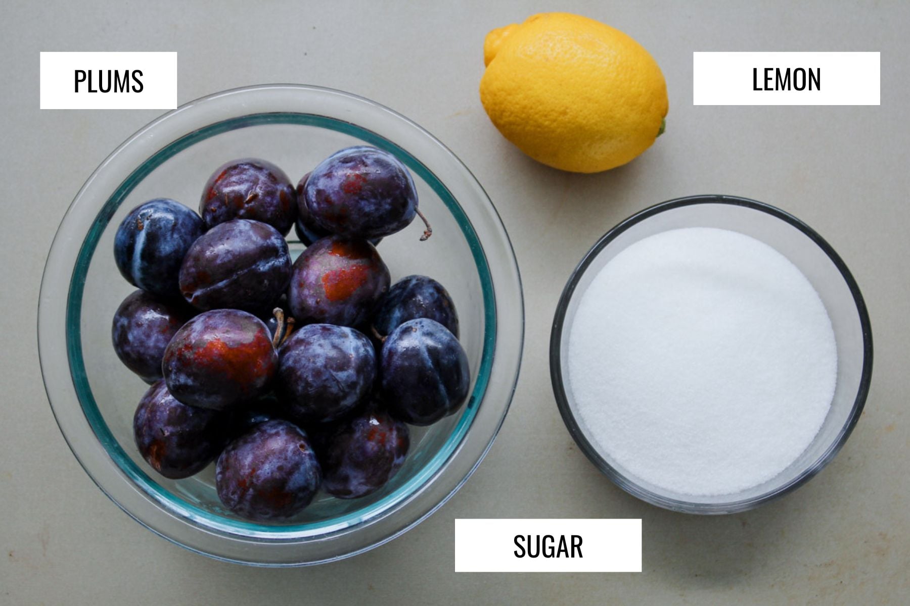 A bowl of dark purple plums, a whole yellow lemon, and a bowl of white sugar on a flat surface, each ingredient labeled&mdash;perfect essentials for making Italian Plum Jam.