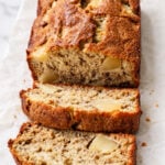 A loaf of Apple Banana Bread with apple chunks rests on parchment paper, with two slices cut and laid in front, showing a moist, textured inside.