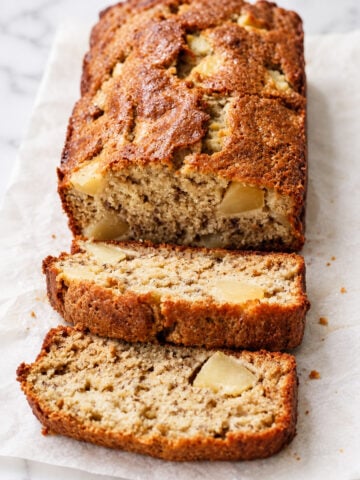 A loaf of Apple Banana Bread with apple chunks rests on parchment paper, with two slices cut and laid in front, showing a moist, textured inside.