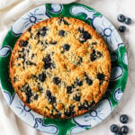 A baked blueberry crumble pie sits on a decorative green and blue plate, reminiscent of a classic Blueberry Lemon Shortbread Tart, with a few fresh blueberries scattered nearby on a white marble surface.