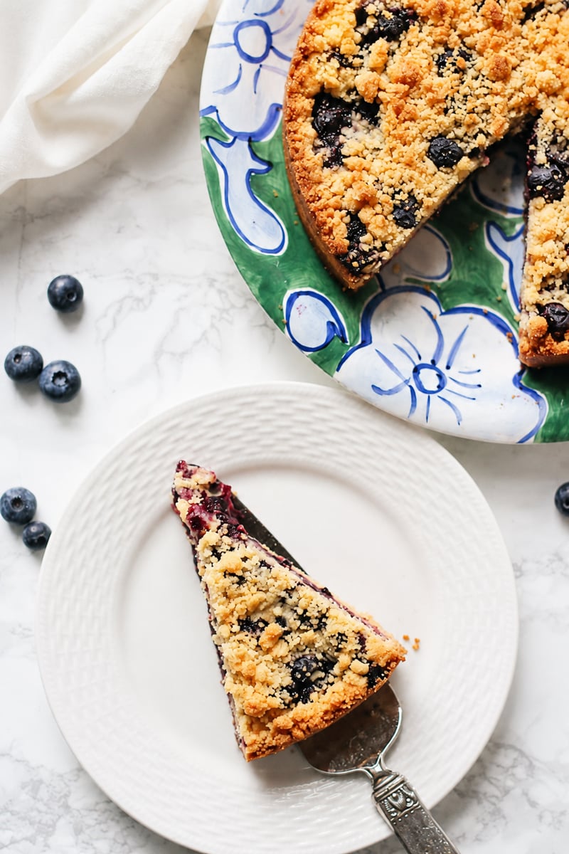 A slice of blueberry crumble cake on a white plate, with the rest of the cake on a colorful floral platter nearby. Fresh blueberries are scattered on the marble surface, evoking all the vibrant flavors of a Blueberry Lemon Shortbread Tart.