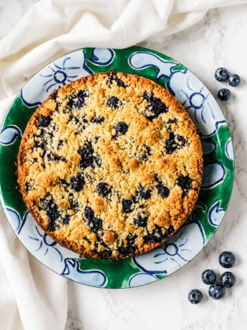 A baked blueberry crumble pie sits on a decorative green and blue plate, reminiscent of a classic Blueberry Lemon Shortbread Tart, with a few fresh blueberries scattered nearby on a white marble surface.