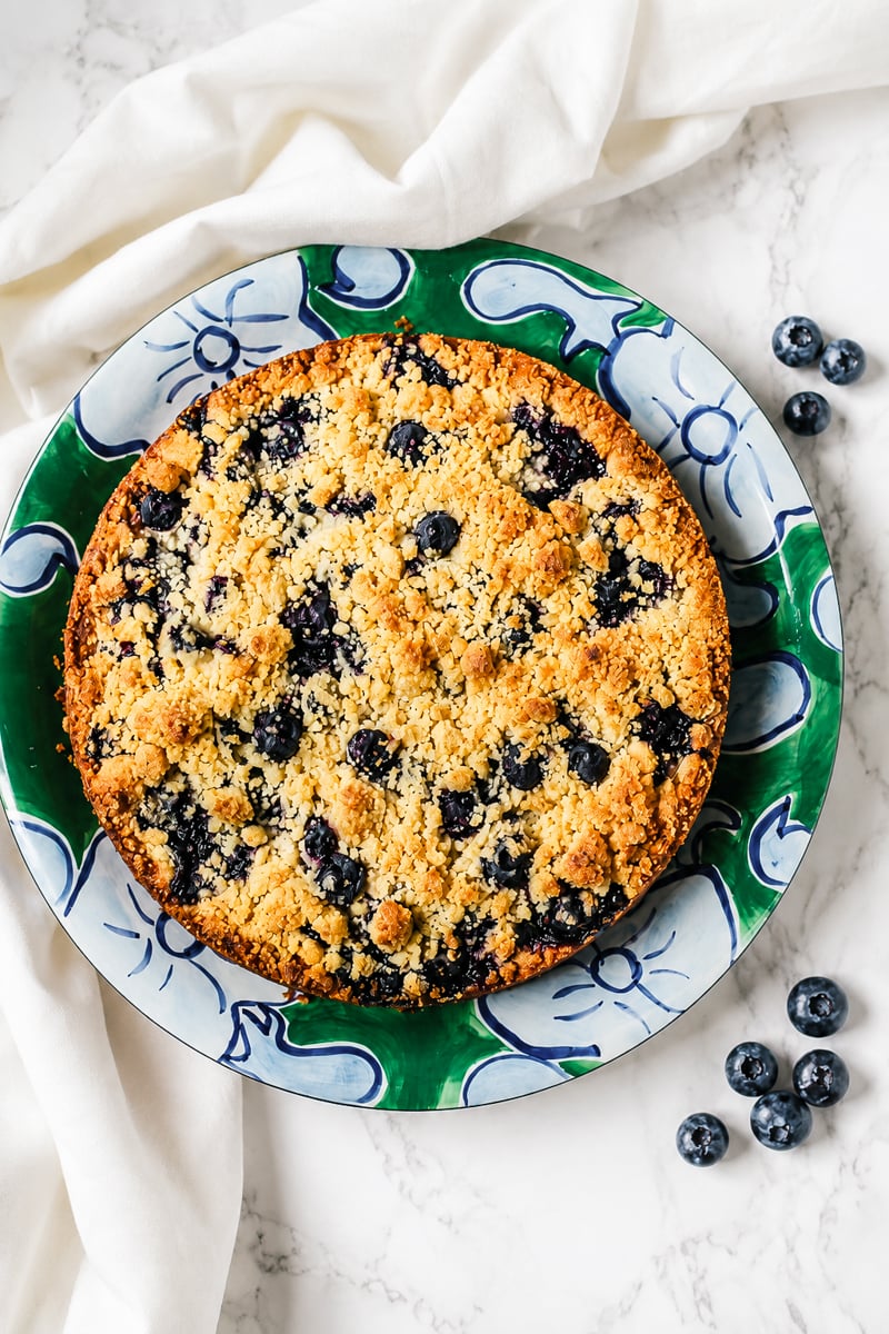 A baked blueberry crumble pie sits on a decorative green and blue plate, reminiscent of a classic Blueberry Lemon Shortbread Tart, with a few fresh blueberries scattered nearby on a white marble surface.