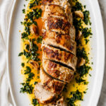 Sliced Rosemary and Garlic Pork Tenderloin on a white platter, surrounded by chopped herbs, roasted garlic cloves, and a drizzle of golden sauce.