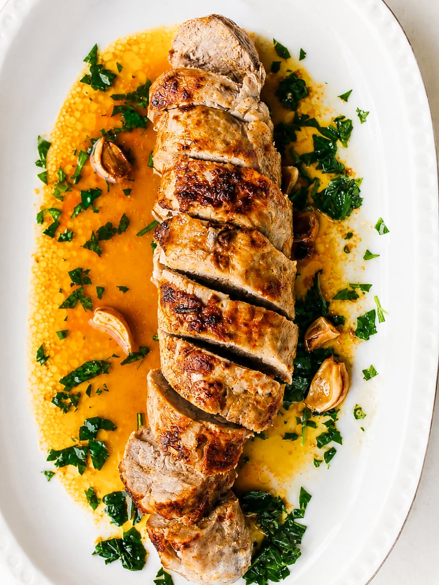 Sliced Rosemary and Garlic Pork Tenderloin on a white platter, surrounded by garlic cloves, chopped parsley, and a golden sauce.