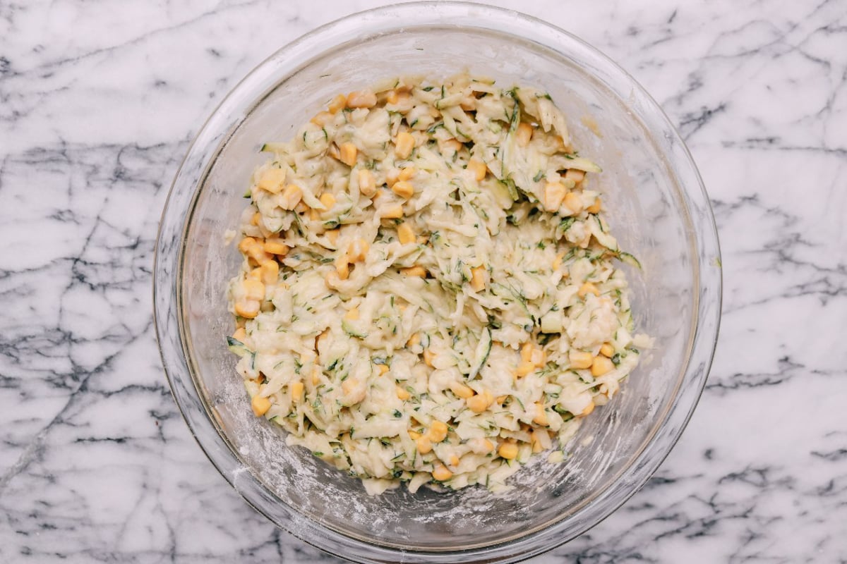 A glass bowl on a marble surface contains the mixture for Zucchini and Corn Fritters-shredded zucchini, corn kernels, and batter-all ready to be cooked.