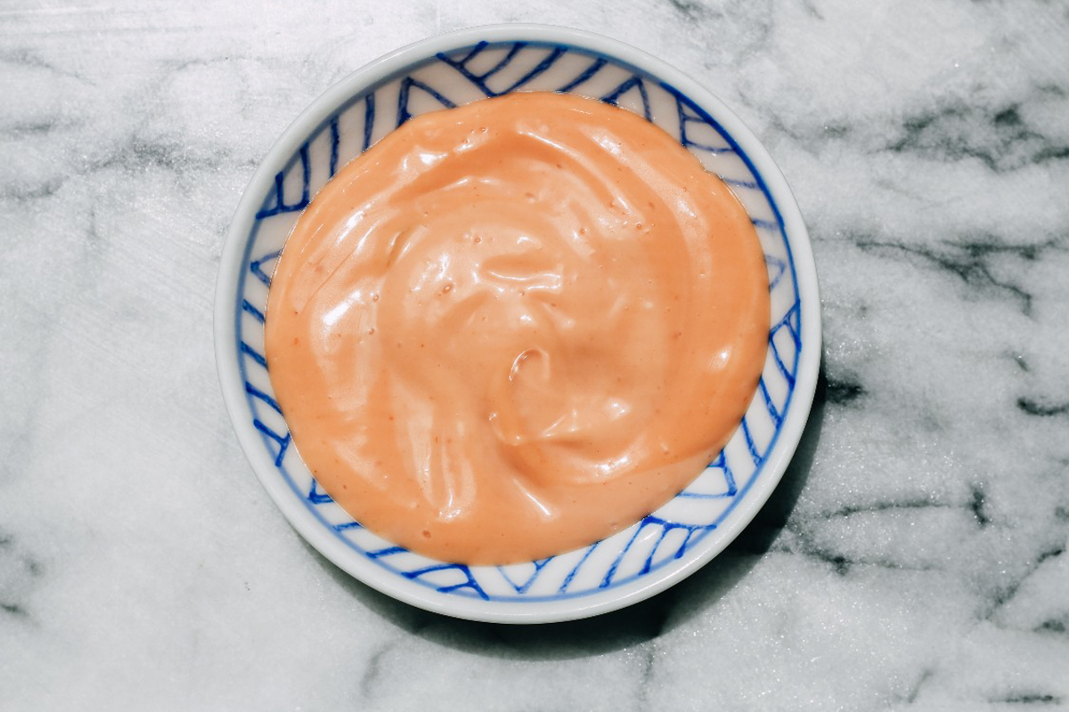 A small round dish with a blue and white pattern, filled with a swirl of pinkish-orange creamy sauce, sits on a marble surface-perfect for dipping zucchini and corn fritters.