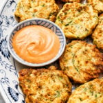A decorative plate holds golden-brown zucchini and corn fritters arranged around a small bowl of creamy dipping sauce. The fritters appear crispy and are speckled with green herbs.