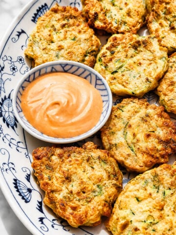 A decorative plate holds golden-brown zucchini and corn fritters arranged around a small bowl of creamy dipping sauce. The fritters appear crispy and are speckled with green herbs.