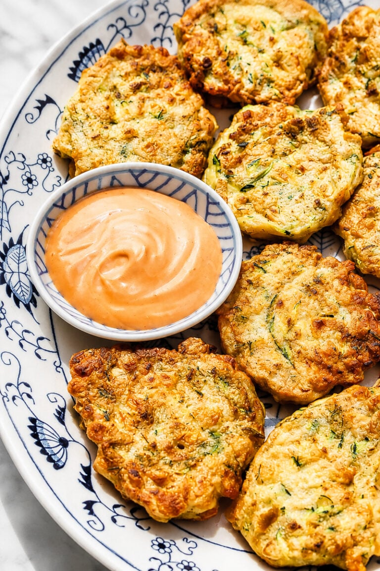 A decorative plate holds golden-brown zucchini and corn fritters arranged around a small bowl of creamy dipping sauce. The fritters appear crispy and are speckled with green herbs.