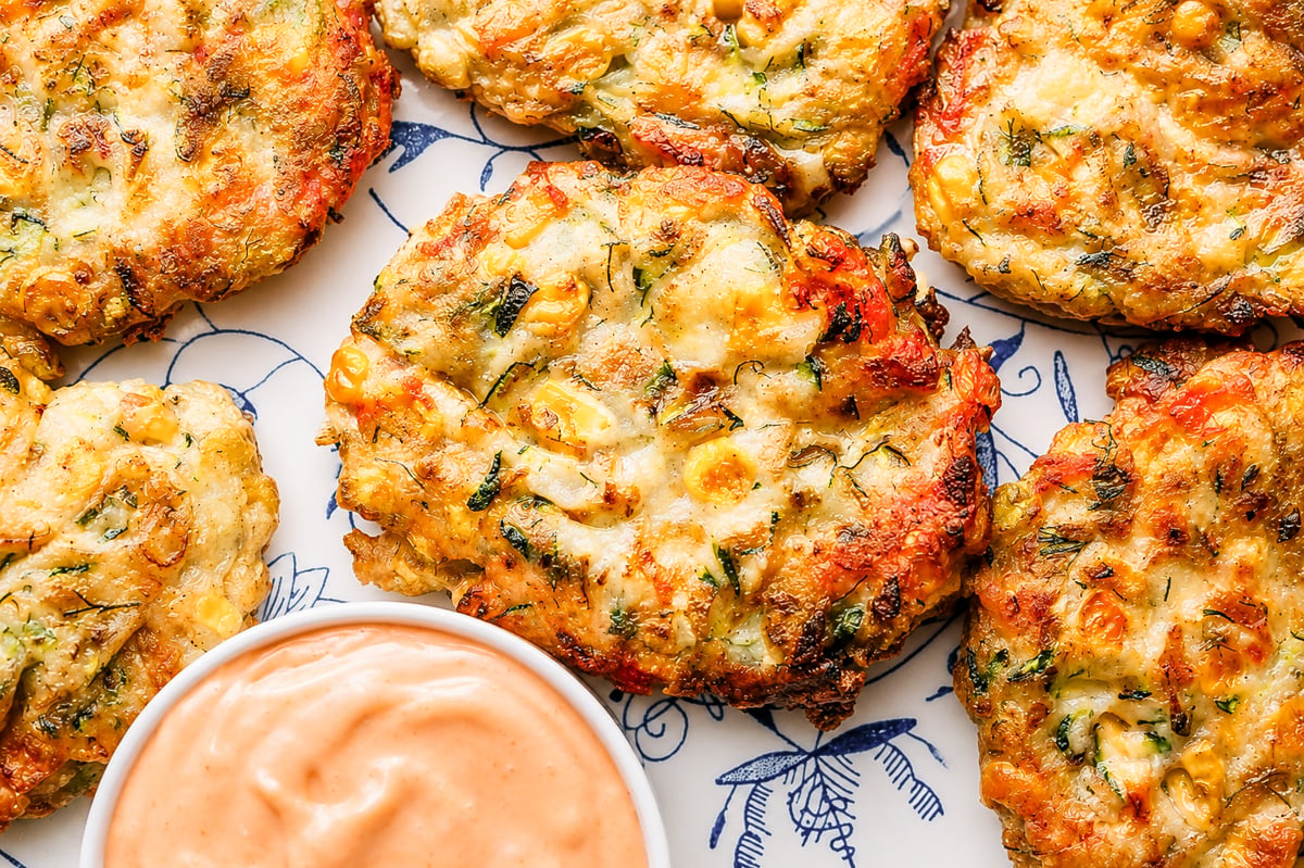 Golden, crispy Zucchini and Corn Fritters are arranged on a patterned plate next to a small bowl of creamy dipping sauce. The fritters show flecks of green herbs and vegetables throughout.