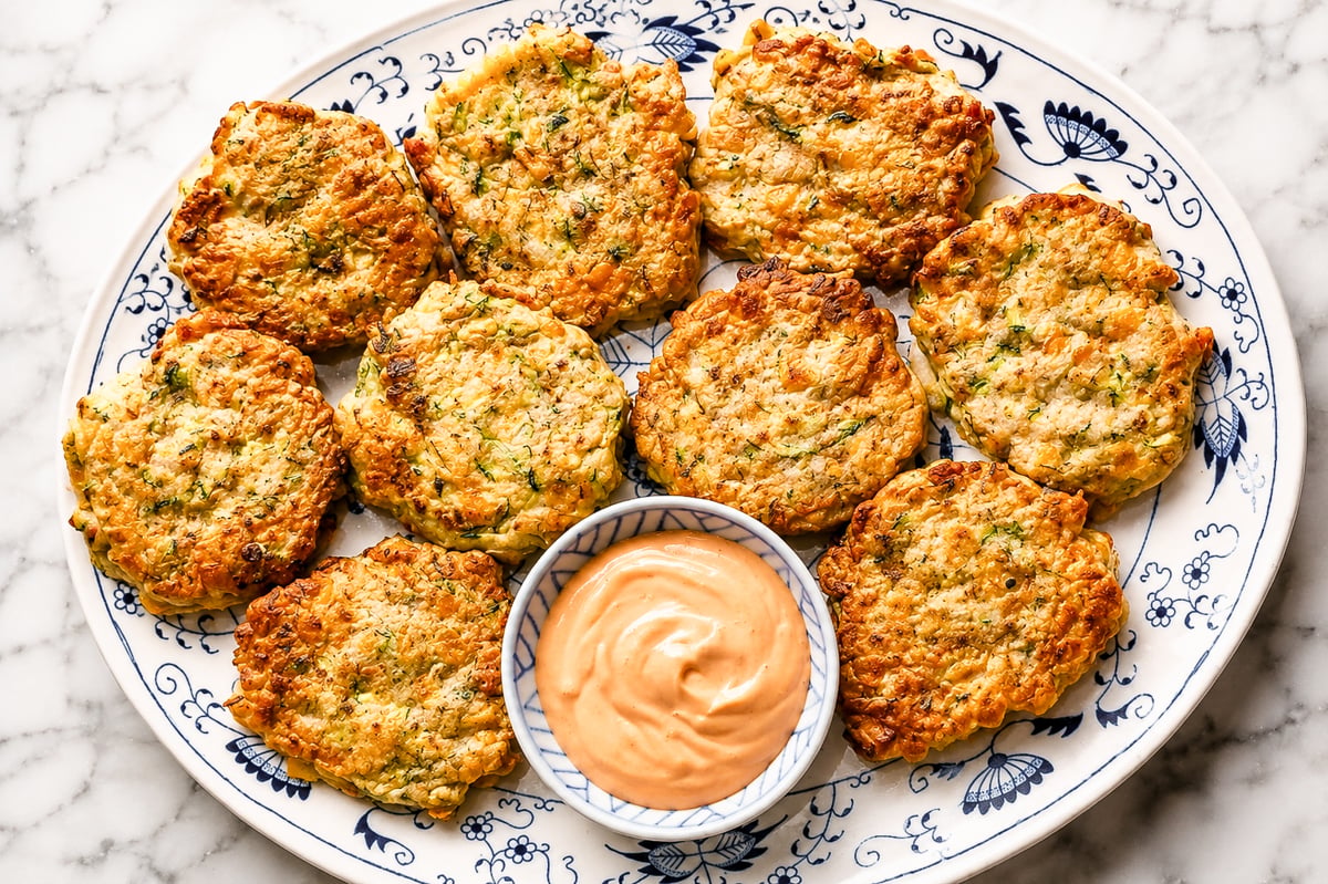A decorative plate holds ten golden-brown zucchini and corn fritters arranged in a circle around a small bowl of creamy orange dipping sauce, all set on a marble surface.